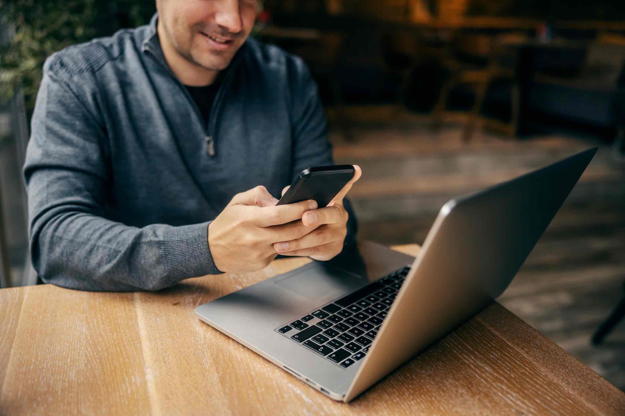 A man, sitting at a table with his laptop, looks at his phone. Social media hacking can open the door to cybersecurity risk.