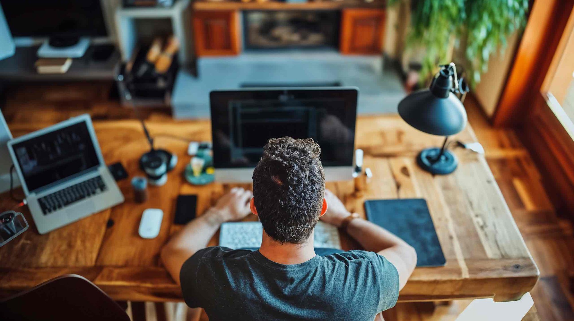 High, behind view of a man working at a large wooden desk. He has a laptop and extra screen.