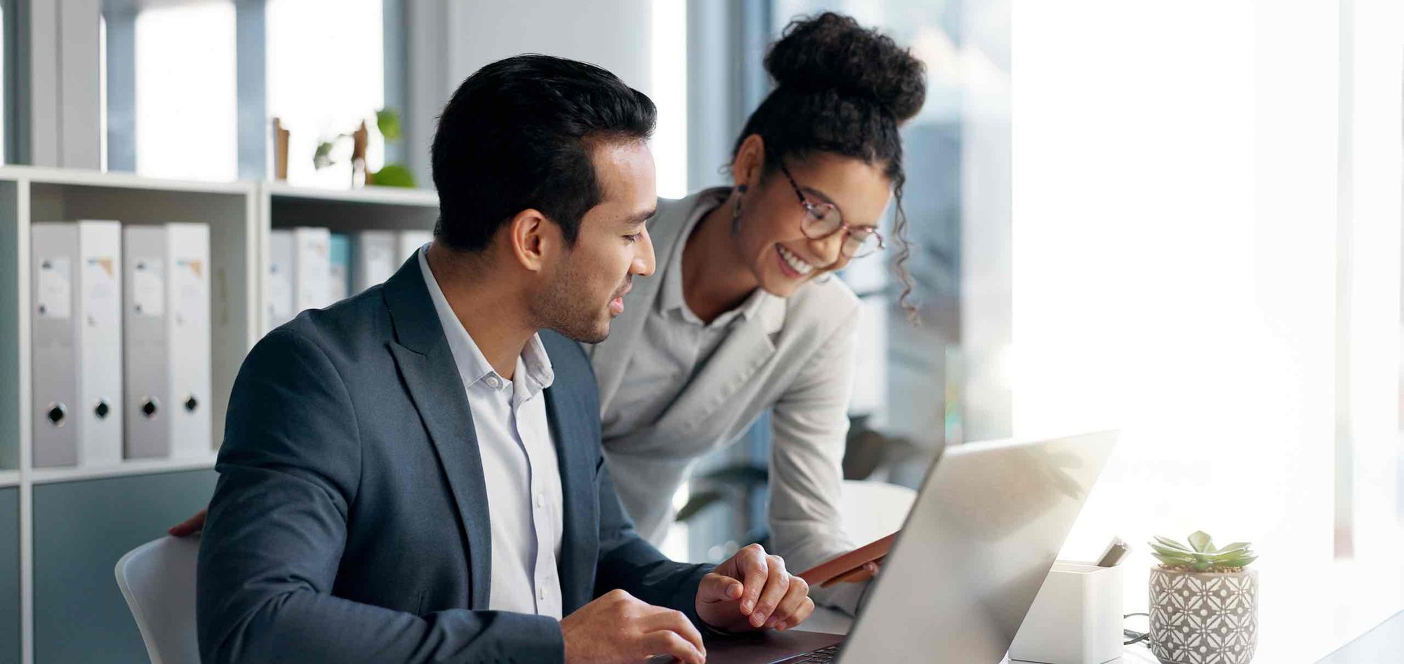 A businesswoman leans over the shoulder of a seated businessman to show him something on her phone.