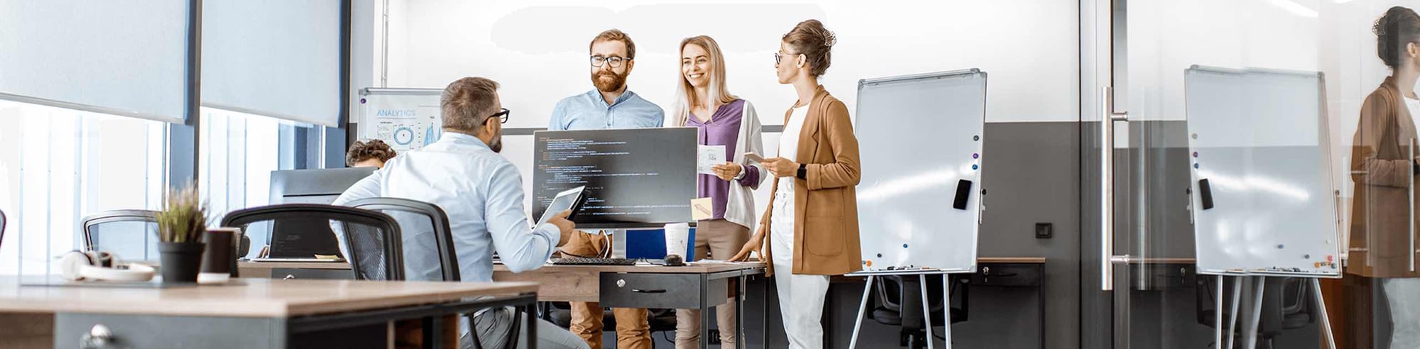 Three workers stand at a colleague's desk and speak to him.