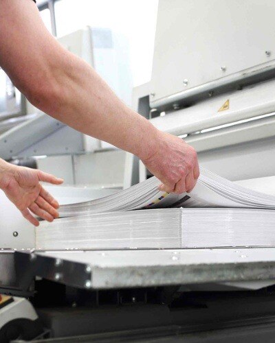 Close-up of hands checking a pile of A3 printouts in a factory setting.