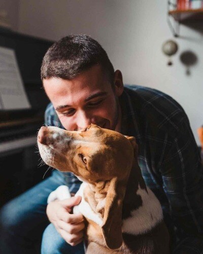 A man and his dog sit at a piano. The man is joyful as he hugs and kisses his dog.