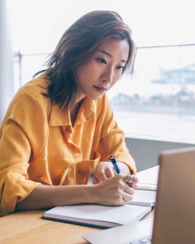 Close-up of an Asian woman in a yellow shirt writing in a notebook while she looks at a laptop screen.