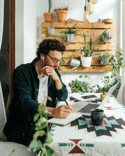 A man working from home with his laptop and notebook. The room is full of pot plants.