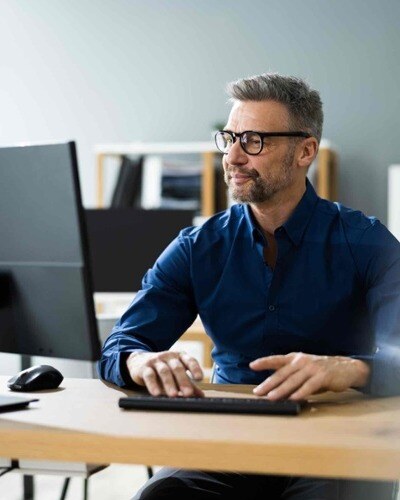 A man in a blue shirt sits at a computer. He appears content.