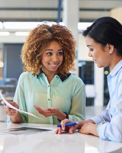 Two women discuss a paper document in a covered, outdoor setting. On-boarding and off-boarding processes are essential.