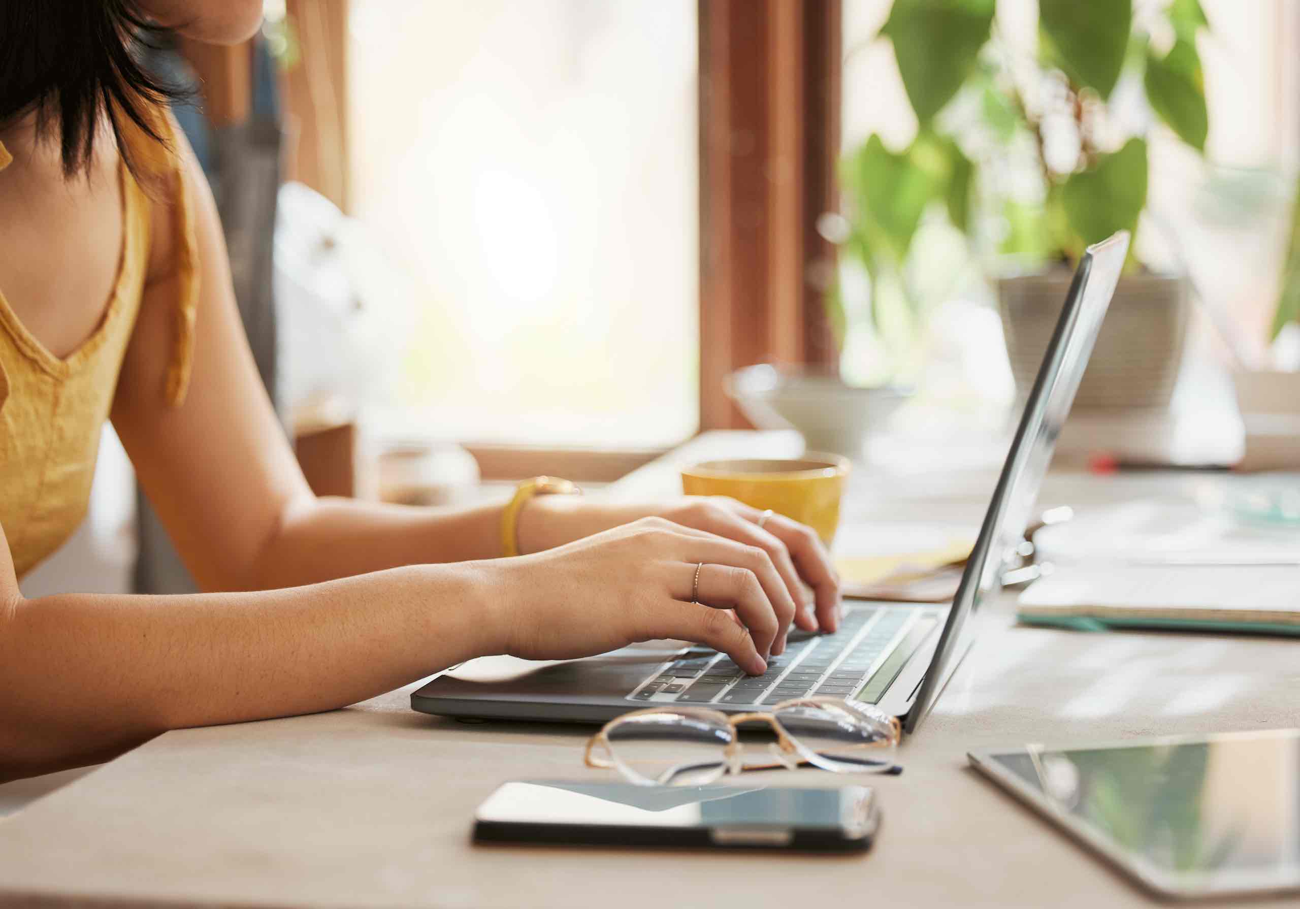 Close-up of a woman's hands typing at a laptop. Glasses and a mobile are in the foreground.