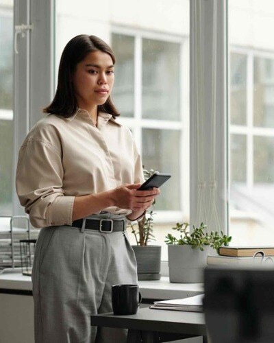 A woman stands near a window in an office environment. She is holding her phone but looking away.