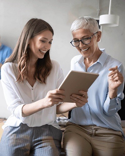 A young woman shows an older woman something on a tablet. Both are laughing.